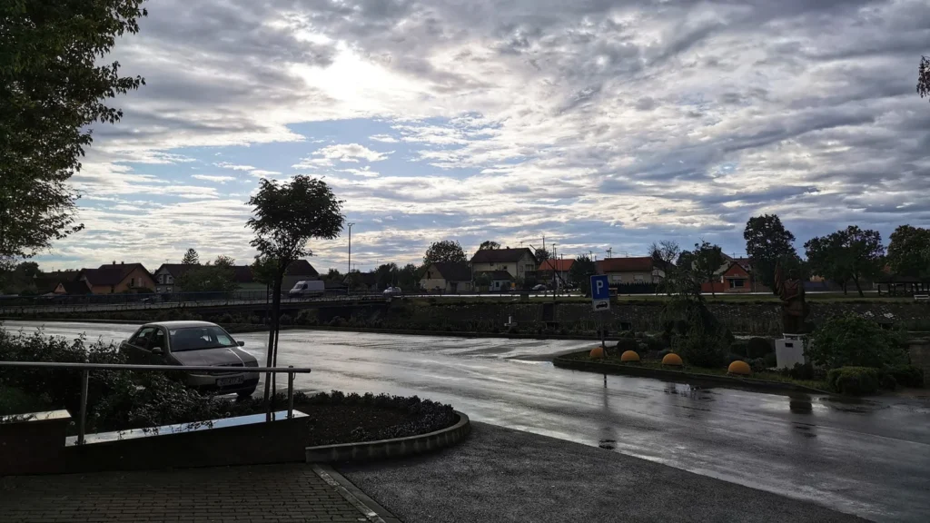Mokra cesta i parkiralište u Cerni pod oblačnim nebom, u pozadini kuće i drveće