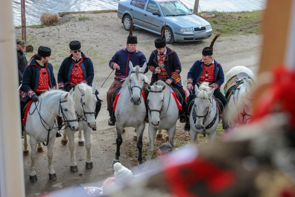 Pet jahača sa šubarama i fazanskim perjem na bijelim konjima ispred kuće uz vodu, na posluženju domaćina