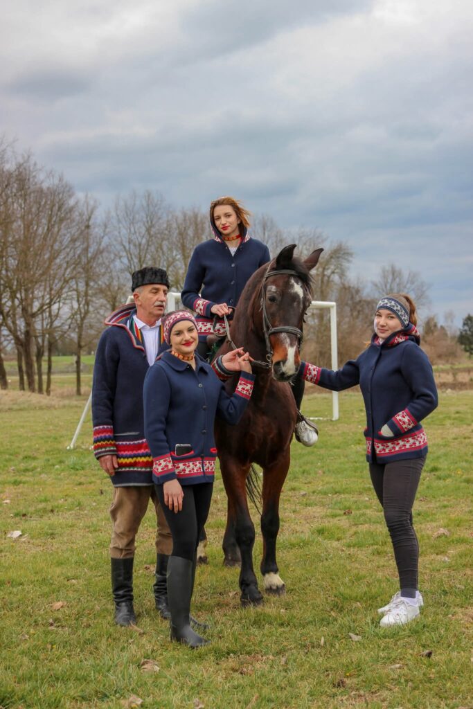Djevojka na konju i troje jahača u tradicijskim kaputima s vezom pozira na livadi uoči pokladnog jahanja